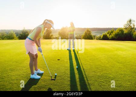 Volle Seitenansicht einer Golfspielerin, die bereit ist, den Ball in den Cup zu schlagen, unter der Anleitung eines qualifizierten Trainers während des Kurses im Freien Stockfoto