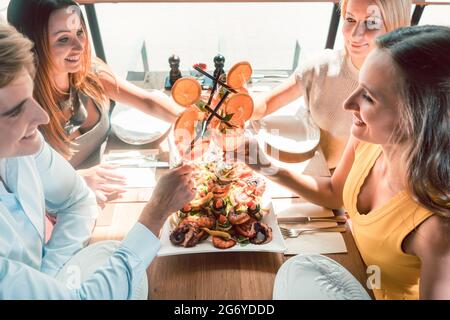 Genießen Sie den Blick auf die Hände von vier jungen Freunden, die mit frischen Orangencocktails toasten, bevor Sie in einem trendigen Restaurant Meeresfrüchte essen Stockfoto