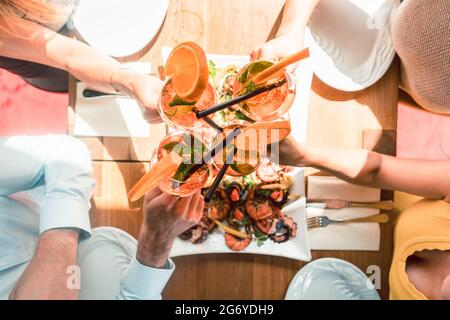 Genießen Sie den Blick auf die Hände von vier jungen Freunden, die mit frischen Orangencocktails toasten, bevor Sie in einem trendigen Restaurant Meeresfrüchte essen Stockfoto