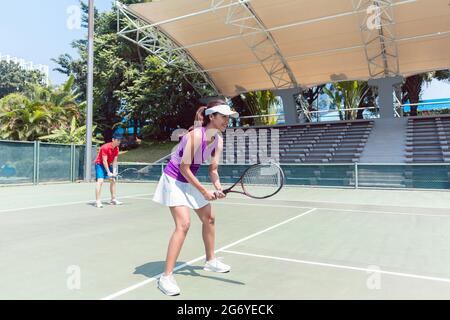 Die volle Länge einer selbstbewussten Tennisspielerin lächelt, während sie darauf wartet, den Ball während des Doppelspieles auf einem professionellen modernen Platz zu treffen Stockfoto