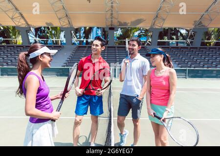 Vier junge und fröhliche Freunde mit einem gesunden Lebensstil sprechen, bevor sie an einem sonnigen Tag ein Doppel-Mixed-Match auf einem professionellen Tennisplatz starten Stockfoto