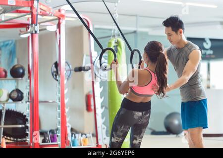 Aus der unteren Perspektive sehen Sie eine schöne junge Frau, die lächelt, während sie während des Trainings im Fitnessstudio Liegestütze auf Gymnastikringen macht Stockfoto