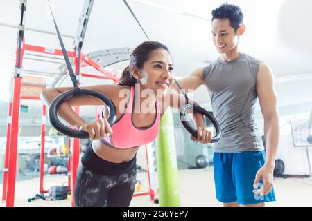 Aus der unteren Perspektive sehen Sie eine schöne junge Frau, die lächelt, während sie Liegestütze auf Gymnastikringen während des funktionellen Trainingstrainings im Fitnessstudio macht Stockfoto