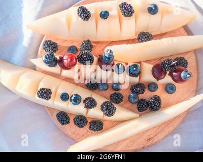 Holzschneidebrett mit frischen und saftigen Brombeeren und Heidelbeeren auf der Oberseite der Melonenscheiben verstreut. Saisonales gesundes Lebensmittelkonzept. Stockfoto