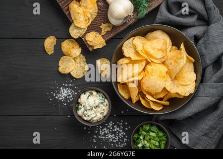 Chips in einer schwarzen Schüssel auf einem Hintergrund aus Gemüse, Sauce und Salz auf einem schwarzen Holztisch. Vorspeise für Bier. Stockfoto