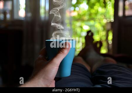 Blaue Tasse Tee raucht in der Hand eines Mannes im Bett. Mann liegt und entspannt an einem heißen Sommertag. Verschwommener Hintergrund der offenen Tür und Blick auf den Garten Stockfoto