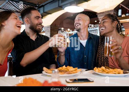 Eine Gruppe verschiedener Freunde, die ihre Biergläser anklimmten, während sie gemeinsam in einem Restaurant essen. Friends-Konzept. Stockfoto