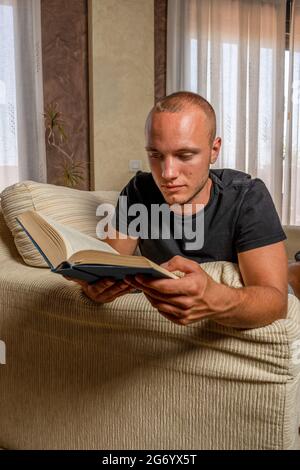 Junger Mann, der sich bequem auf der Couch ausdehnt und ein Buch liest. Mann, der sich zu Hause entspannt. Kulturkonzept Stockfoto