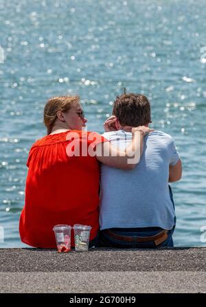 Junge Liebespaare, die am Meer kuscheln, sitzen an einer Wand mit Armen umeinander. Stockfoto