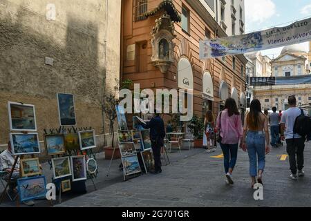 Straßenkünstler verkaufen ihre Gemälde in der Via San Lorenzo im historischen Zentrum von Genua mit Touristen, die im Sommer, Ligurien, Italien, spazieren Stockfoto