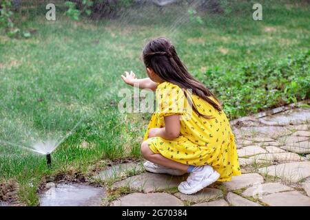 Rückansicht eines Mädchens, das mit Wassertropfen aus einem Rasensprenger spielt, hilft ein Mädchen ihren Eltern, das Gras auf dem Rasen zu bewässern Stockfoto