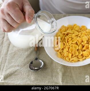 Der beschnittene Mann gießt Milch in Cornflakes auf einem weißen Teller auf einer schlichten, rauen Tischdecke. Blick von oben. Selektiver Fokus. Konzept, einfach schnell amerikanischen h Stockfoto