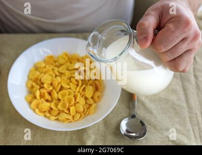 Der beschnittene Mann gießt Milch in Cornflakes auf einem weißen Teller auf einer schlichten, rauen Tischdecke. Blick von oben. Selektiver Fokus. Konzept, einfach schnell amerikanischen h Stockfoto