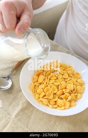 Der beschnittene Mann gießt Milch in Cornflakes auf einem weißen Teller auf einer schlichten, rauen Tischdecke. Blick von oben. Selektiver Fokus. Konzept, einfach schnell amerikanischen h Stockfoto