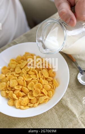 Der beschnittene Mann gießt Milch in Cornflakes auf einem weißen Teller auf einer schlichten, rauen Tischdecke. Blick von oben. Selektiver Fokus. Konzept, einfach schnell amerikanischen h Stockfoto