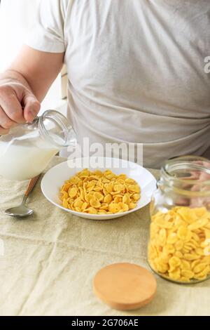 Der beschnittene Mann gießt Milch in Cornflakes auf einem weißen Teller auf einer schlichten, rauen Tischdecke. Blick von oben. Selektiver Fokus. Konzept, einfach schnell amerikanischen h Stockfoto