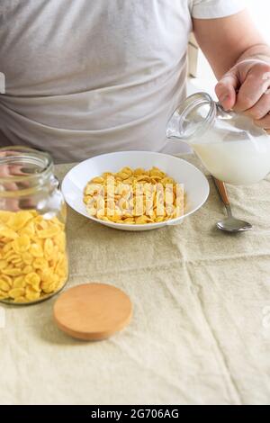 Der beschnittene Mann gießt Milch in Cornflakes auf einem weißen Teller auf einer schlichten, rauen Tischdecke. Blick von oben. Selektiver Fokus. Konzept, einfach schnell amerikanischen h Stockfoto