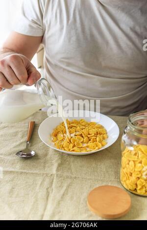 Der beschnittene Mann gießt Milch in Cornflakes auf einem weißen Teller auf einer schlichten, rauen Tischdecke. Blick von oben. Selektiver Fokus. Konzept, einfach schnell amerikanischen h Stockfoto