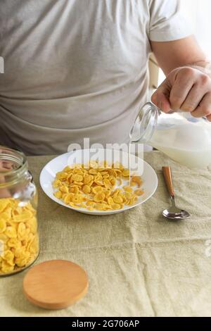 Der beschnittene Mann gießt Milch in Cornflakes auf einem weißen Teller auf einer schlichten, rauen Tischdecke. Blick von oben. Selektiver Fokus. Konzept, einfach schnell amerikanischen h Stockfoto