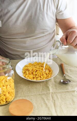 Der beschnittene Mann gießt Milch in Cornflakes auf einem weißen Teller auf einer schlichten, rauen Tischdecke. Blick von oben. Selektiver Fokus. Konzept, einfach schnell amerikanischen h Stockfoto