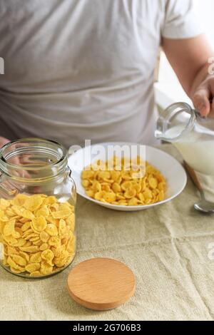 Der beschnittene Mann gießt Milch in Cornflakes auf einem weißen Teller auf einer schlichten, rauen Tischdecke. Blick von oben. Selektiver Fokus. Konzept, einfach schnell amerikanischen h Stockfoto