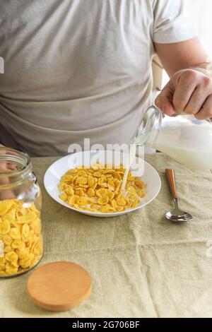 Der beschnittene Mann gießt Milch in Cornflakes auf einem weißen Teller auf einer schlichten, rauen Tischdecke. Blick von oben. Selektiver Fokus. Konzept, einfach schnell amerikanischen h Stockfoto