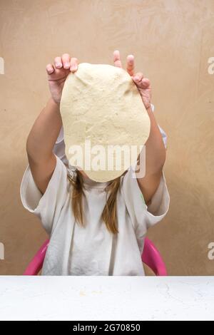 Ein kleines Mädchen hält den Teig nach Gewicht, bedeckt ihr Gesicht. Die Entwicklung der Feinmotorik bei Kindern. Salzteig modellieren. Stockfoto