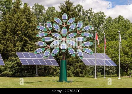 Sonnenkollektoren auf den Blättern einer baumförmigen Metallstruktur. Drei Sonnenkollektoren und Flaggen, darunter die kanadische Flagge, im Hintergrund. Stockfoto