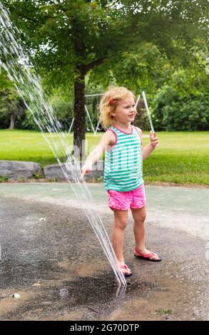 Kleine glückliche Kinder, die am heißen, sonnigen Sommertag auf dem Splash Pad im öffentlichen Park mit Wasser spielen. Kleine, schöne Kinder, die sich am Springbrunnen amüsieren Stockfoto