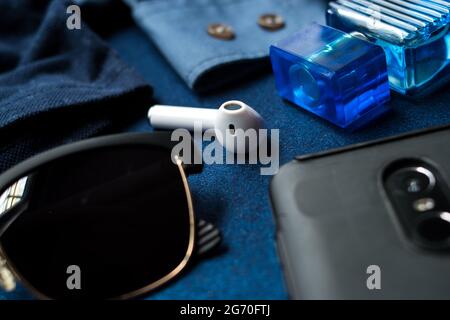 Einige Sonnenbrillen, schwarzes Handy, kabelloser Kopfhörer und Glaslotionflasche auf einem blau strukturierten Schreibtisch Stockfoto