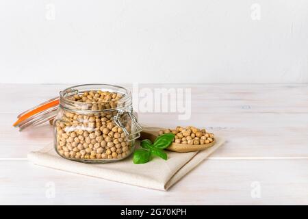 Rohe Kichererbsen in einem Glas mit Cliptop und in einem großen Holzlöffel auf einem weißen rustikalen Tisch. Trockene Kichererbsen für Hummus. Pflanzliches Eiweiß aus Hülsenfrüchte. Stockfoto