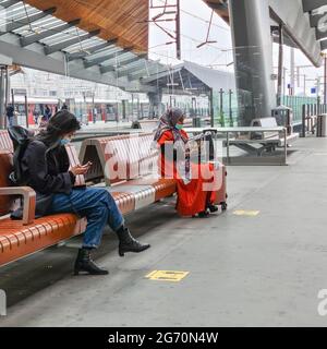 Amsterdam, Niederlande, 25. Juni 2021 : zwei Damen unterschiedlicher Kulturen, die Gesichtsmasken tragen und ihre Telefone überprüfen, während sie auf den Bänken in der Bahnhofshalle der Amsterdam Bijlmer Arena sitzen und auf ihren Zug warten. Gelbe Markierungen auf dem Boden zeigen eine Entfernung von 1,5 Metern an, eine der Grundregeln während der covid Pandemie. Stockfoto