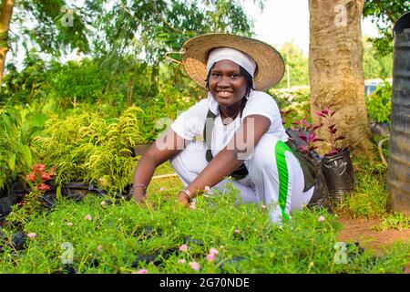 Afrikanische Gärtnerin, Floristin oder Gärtnerin, die eine Schürze und einen Hut trägt, arbeitet während sie in einer grünen und bunten Blumen und Pflanzen gard hockt Stockfoto