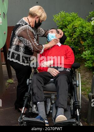Vertikale Aufnahme eines kranken älteren Mannes, der im Rollstuhl neben einer Helferin im Freien eine Maske trägt Stockfoto