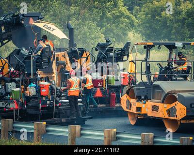 Mehrere Arbeiter mit Harthüten und reflektierenden orangefarbenen Westen betreiben CAT-Asphaltpflastermaschinen und Muldenkipper auf dem Highway 25 in Quebec. Stockfoto