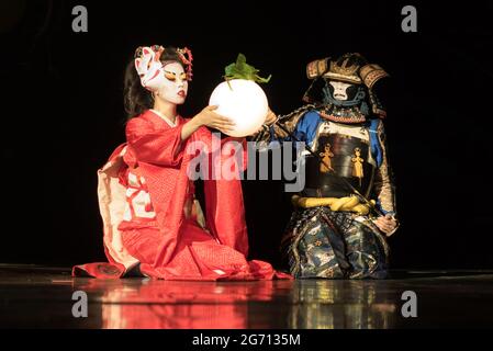 Japanische Geisha in traditionellem Kimono und Fuchsmaske mit Kugellampe und Samurai-Krieger in Rüstung sitzen im Dunkeln auf den Knien. Traditionell Stockfoto