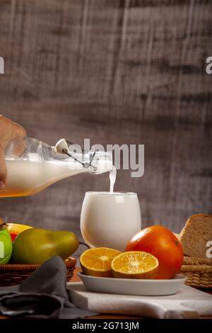 Vertikaler Schuss von Hand gießt frische Milch auf Glas mit Früchten an den Seiten Stockfoto