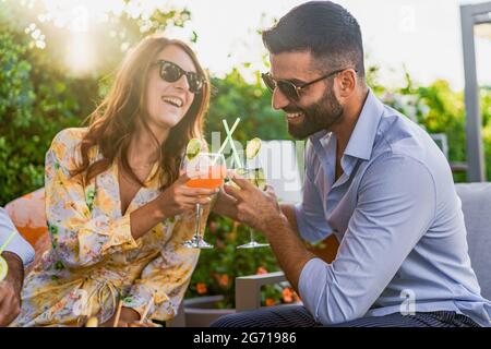 Ein paar junge Freunde, die bei einer Gartenparty auf einem Sofa auf einem festlichen Toast mit klirrenden Gläsern und Fruchtcocktails sitzen. Junge Menschen Stockfoto