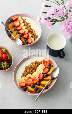 Leckeres gesundes Frühstück, Müsli mit Joghurt und süßen Früchten, Banane und Erdbeeren, Blaubeeren und Pfirsiche, Milch im Becher und Blumen Stockfoto