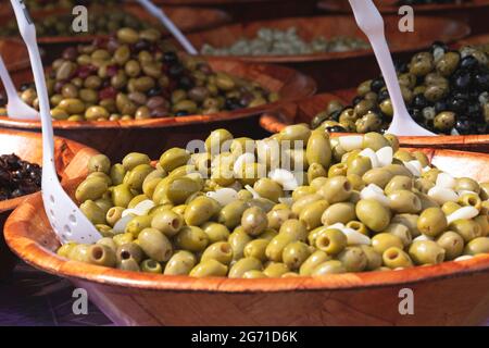 Braune Schale mit grünen Oliven in Olivenöl und weißen Servierlöffeln in den Schalen Stockfoto