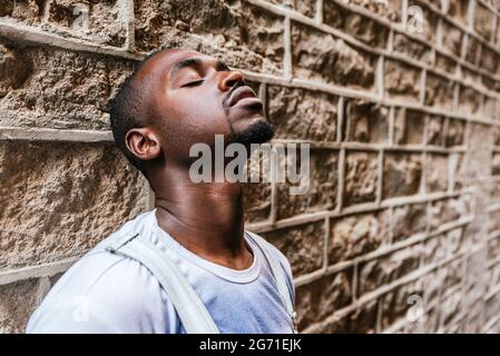 Afro attraktiver Mann, der sich mit geschlossenen Augen an einer Wand lehnt. Konzept der Reflexion, Gedanken, Traurigkeit Stockfoto