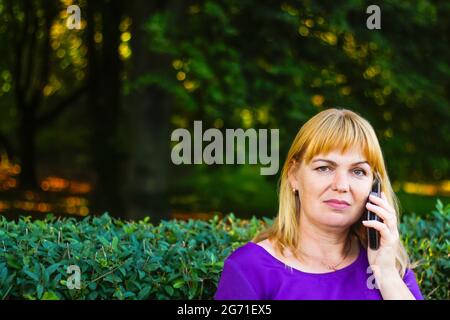 Unschärfe kaukasisch blonde Frau reden, sprechen am Telefon draußen, im Freien. 40-jährige Frau in lila Bluse im Park auf der Bank. Erwachsene, weiblich pro Stockfoto