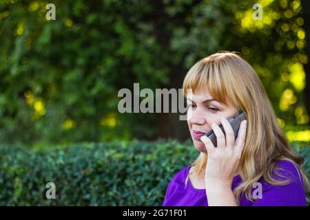 Unschärfe kaukasisch blonde Frau sprechen auf dem Telefon draußen, draußen. 40-jährige Frau in lila Bluse im Park. Seriöse Person mit mobilen, smartp Stockfoto