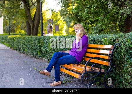 Unschärfe-Eleganz kaukasische blonde Frau reden, sprechen auf dem Telefon draußen, im Freien. 40-jährige Frau in lila Bluse im Park. Erwachsene Frauen usin Stockfoto