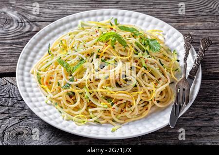 Sommer-Spaghetti alla carrettiera mit Minzblättern, Zitronenschale, Pecorino-Käse, Chilischoten-Flocken, Petersilie und geriebenen Zucchini auf einem weißen Teller Stockfoto