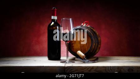 Rotweinglas mit Flasche, Kork und Korkenzieher und kleinem Fass auf rustikalem Holztisch und rotem Hintergrund, Nahaufnahme, Platz für Text. Stockfoto