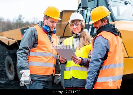 Drei Arbeiter in einem Steinbruch diskutieren vor schweren Maschinen, um Plan zu betrachten Stockfoto