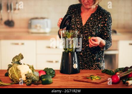 Weibliche Ernte mittleren Alters, die reifes Gemüse in den Mixer geben, während sie in der Küche zu Hause gesunden Smoothie kochen Stockfoto