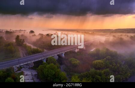 Drohnenansicht der hellen Dämmerung über der Autobahnüberführung, Wald und nebliger Wiese. Stockfoto