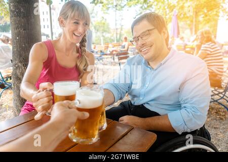 Freunde im Biergarten trinken, ein Mann im Rollstuhl, alle sind anstoßen Stockfoto